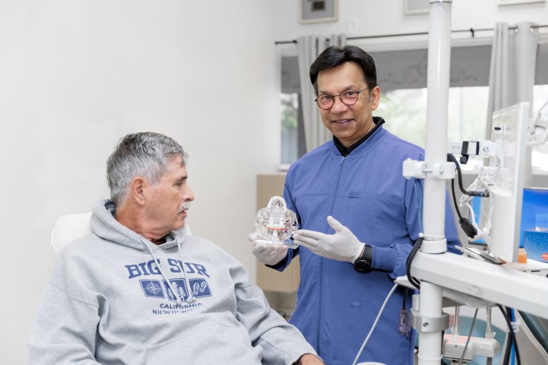 A dentist in blue scrubs and gloves shows a dental model to an older male patient seated in the chair — clinical consultation in a modern operatory